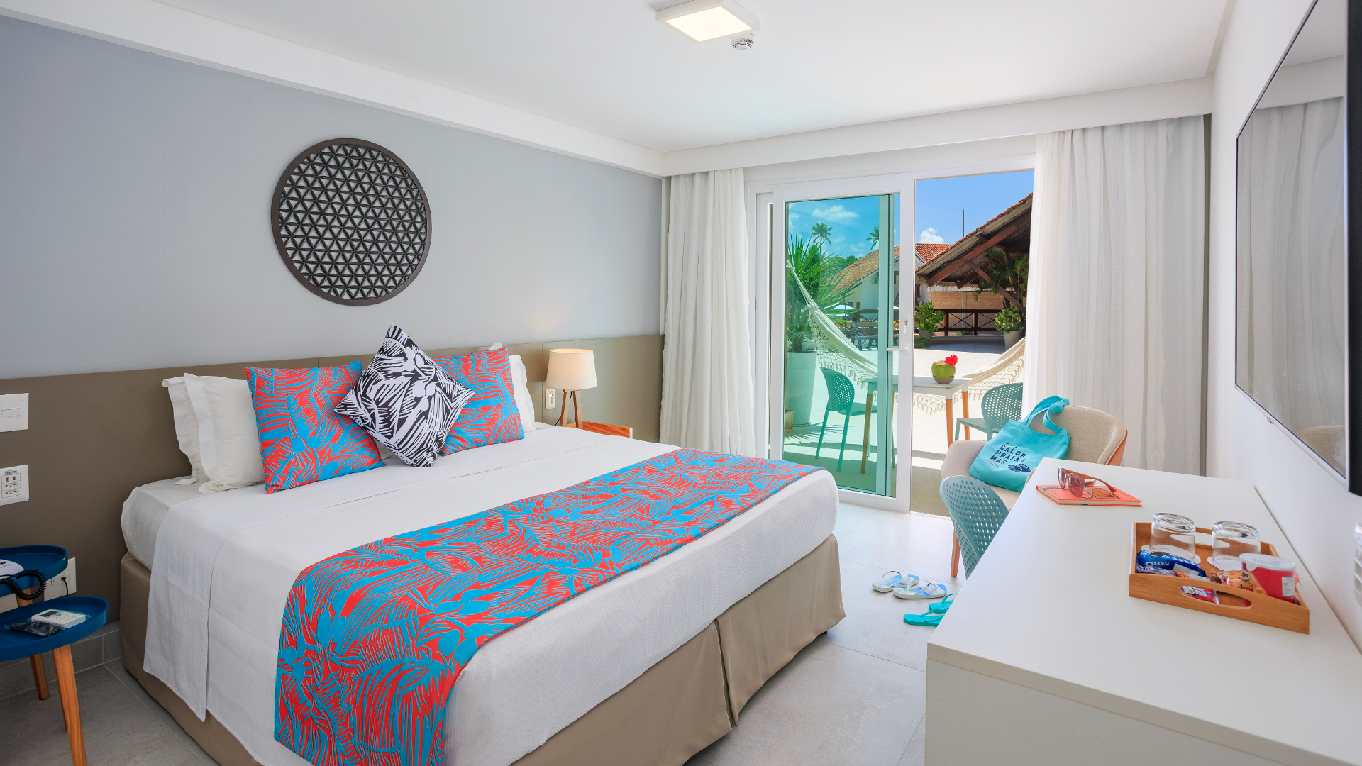 Quarto de hotel iluminado com cama de casal, roupa de cama branca com detalhes em azul e coral, varanda com vista para área externa e decoração moderna em tons neutros.