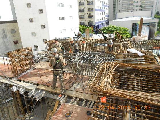 obra caravelle 3 - Conx Construtora Trabalhadores da construção civil construindo com vergalhões no telhado de um arranha-céu. Alguns usam cintos de segurança. Luz do dia intensa.