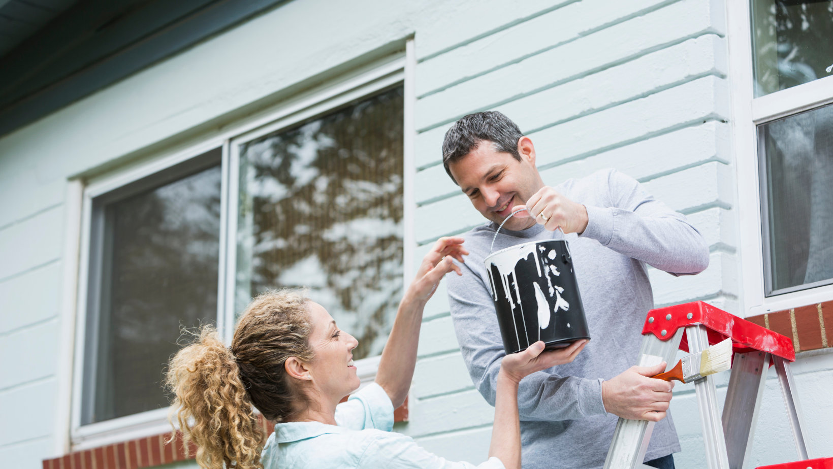 Two people passing a paint can as they paint their home's exterior.