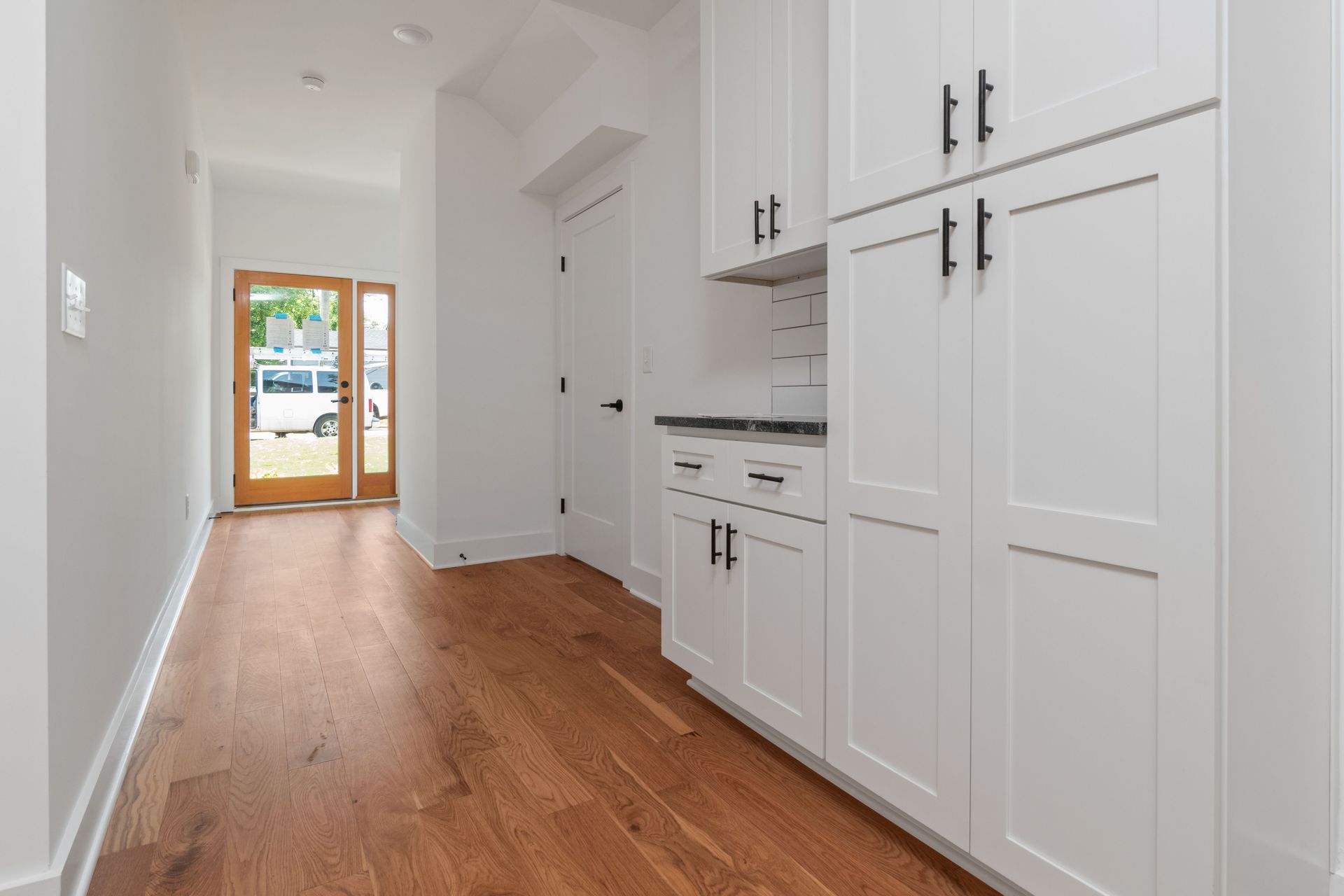 Bright hallway with white cabinets, wood floor, and a doorway to a sunlit room.