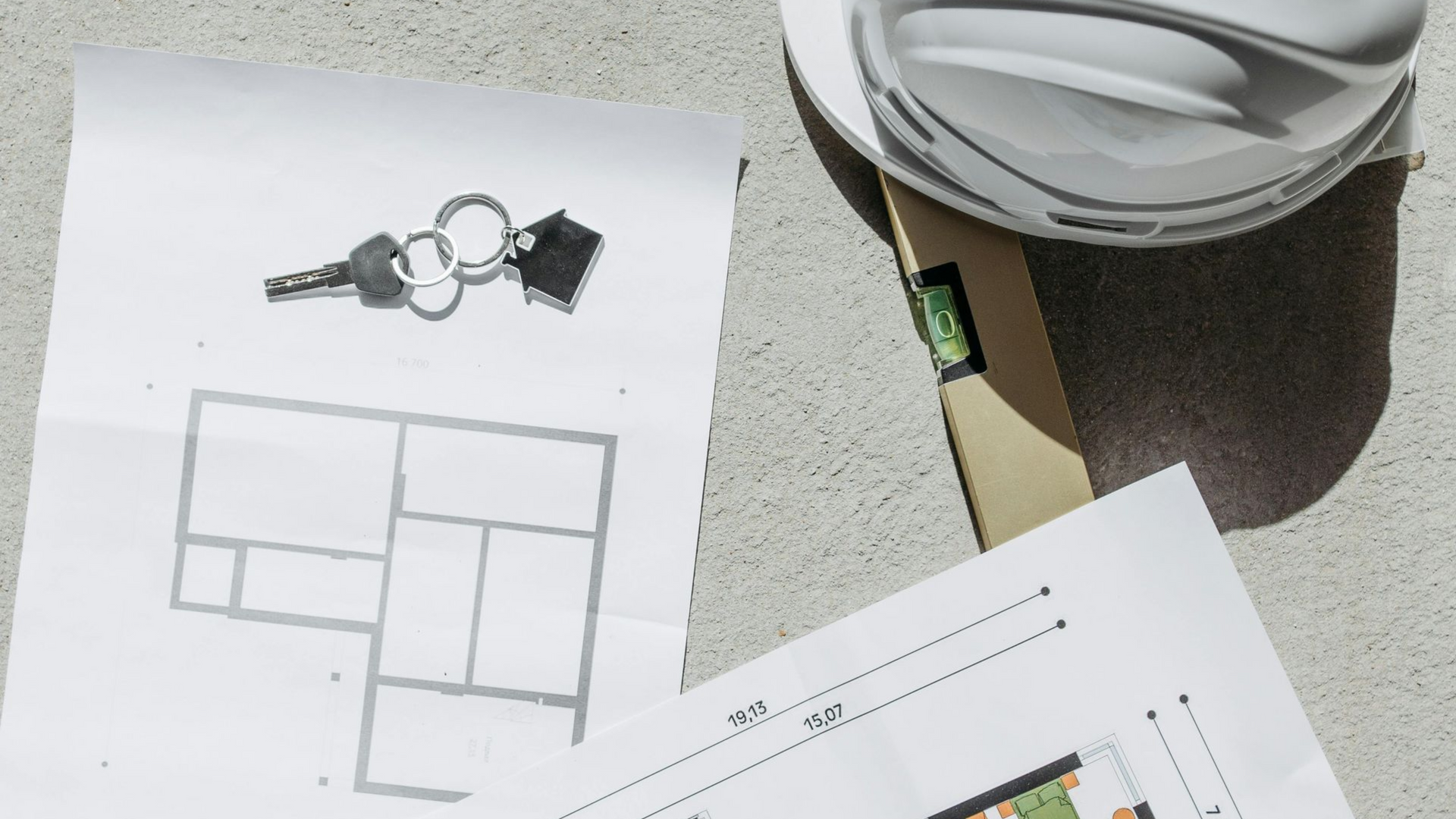 A white hard hat, a wooden level, and two architectural floor plans lay on a grey concrete surface.