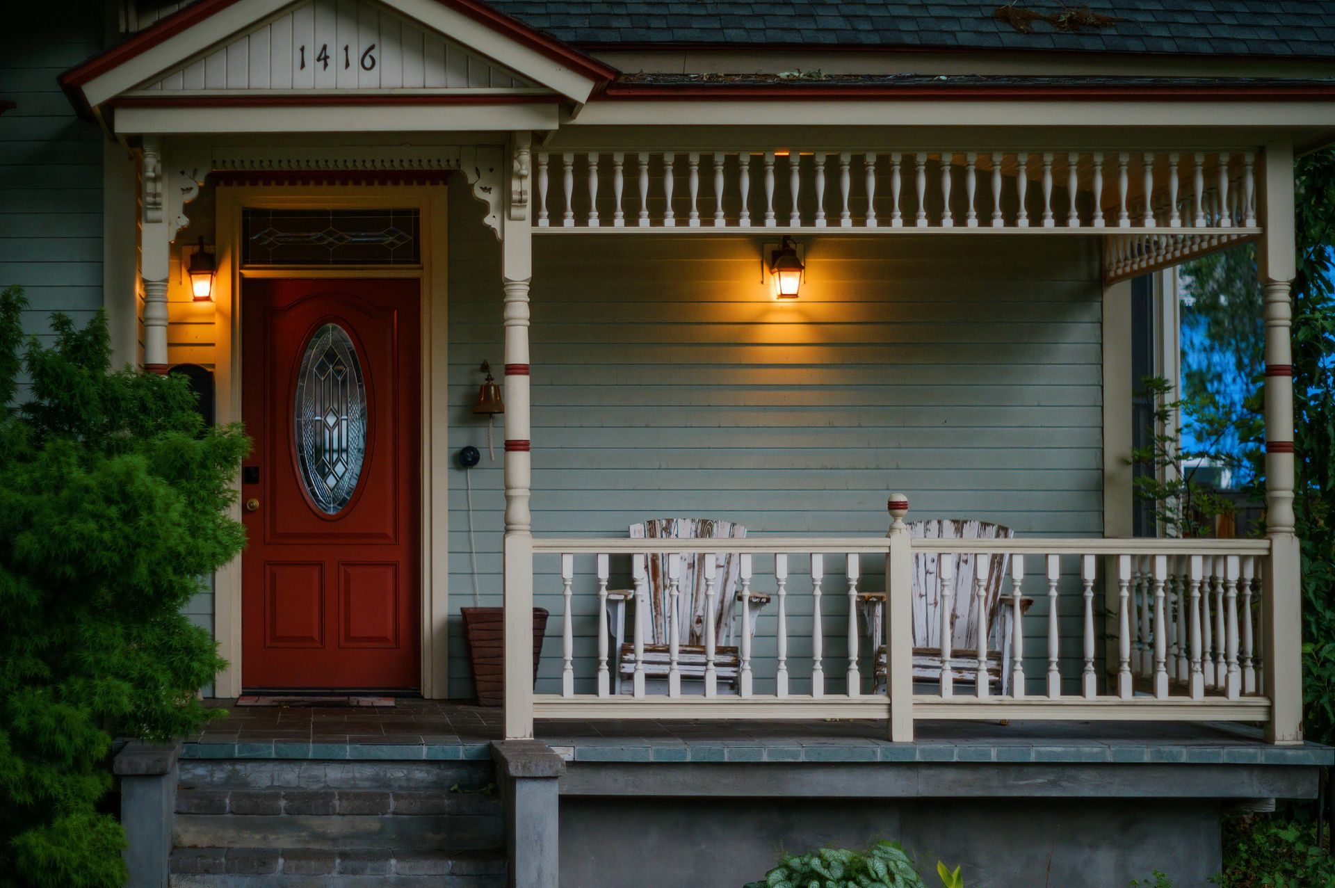 Front porch of a house with light-colored siding, a red door, two chairs, and lit wall lamps. Number 1416 is above the door.