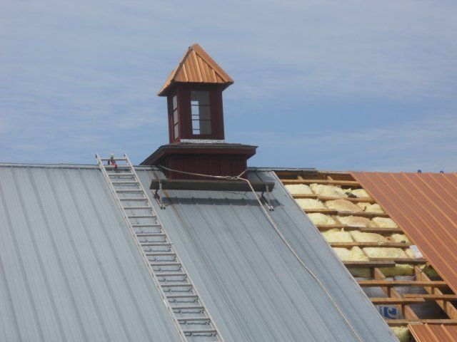 Metal roof installation with a ladder, cupola, and partially exposed roof framing.