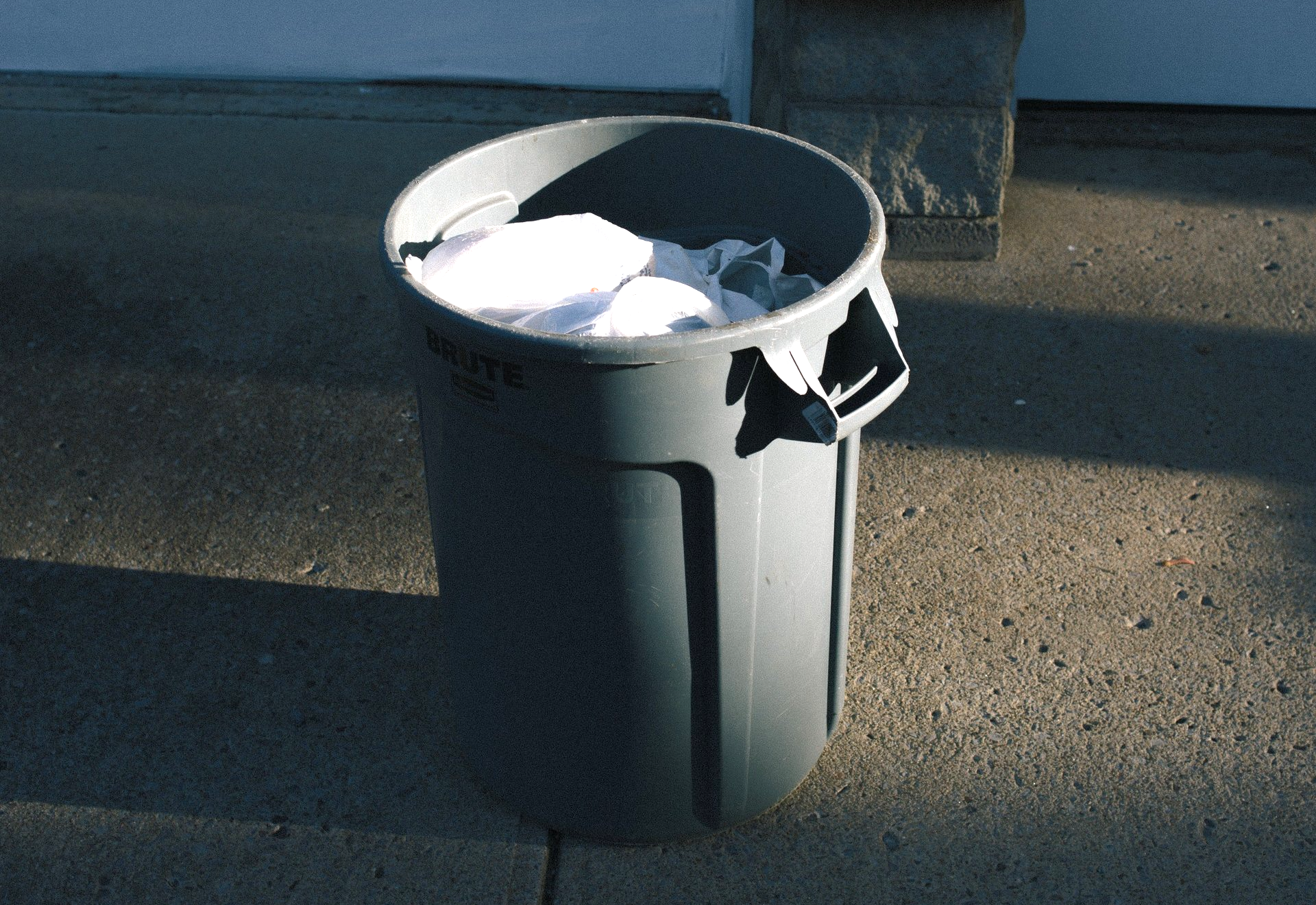 gray garbage can sitting in driveway with a white trash bag inside