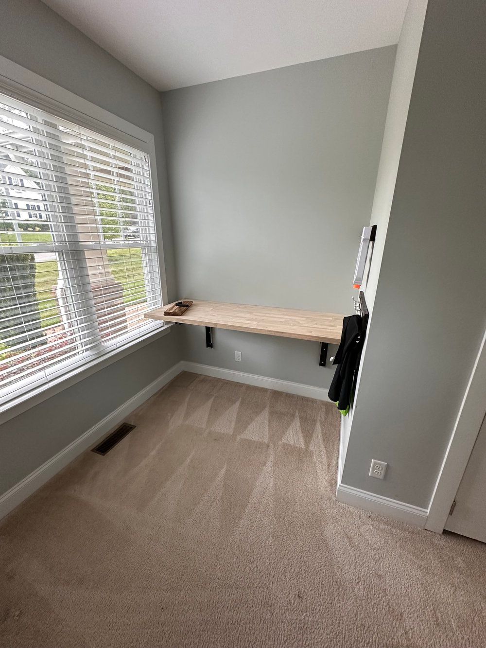 Empty home office corner with a wall-mounted desk, window blinds, and beige carpet.