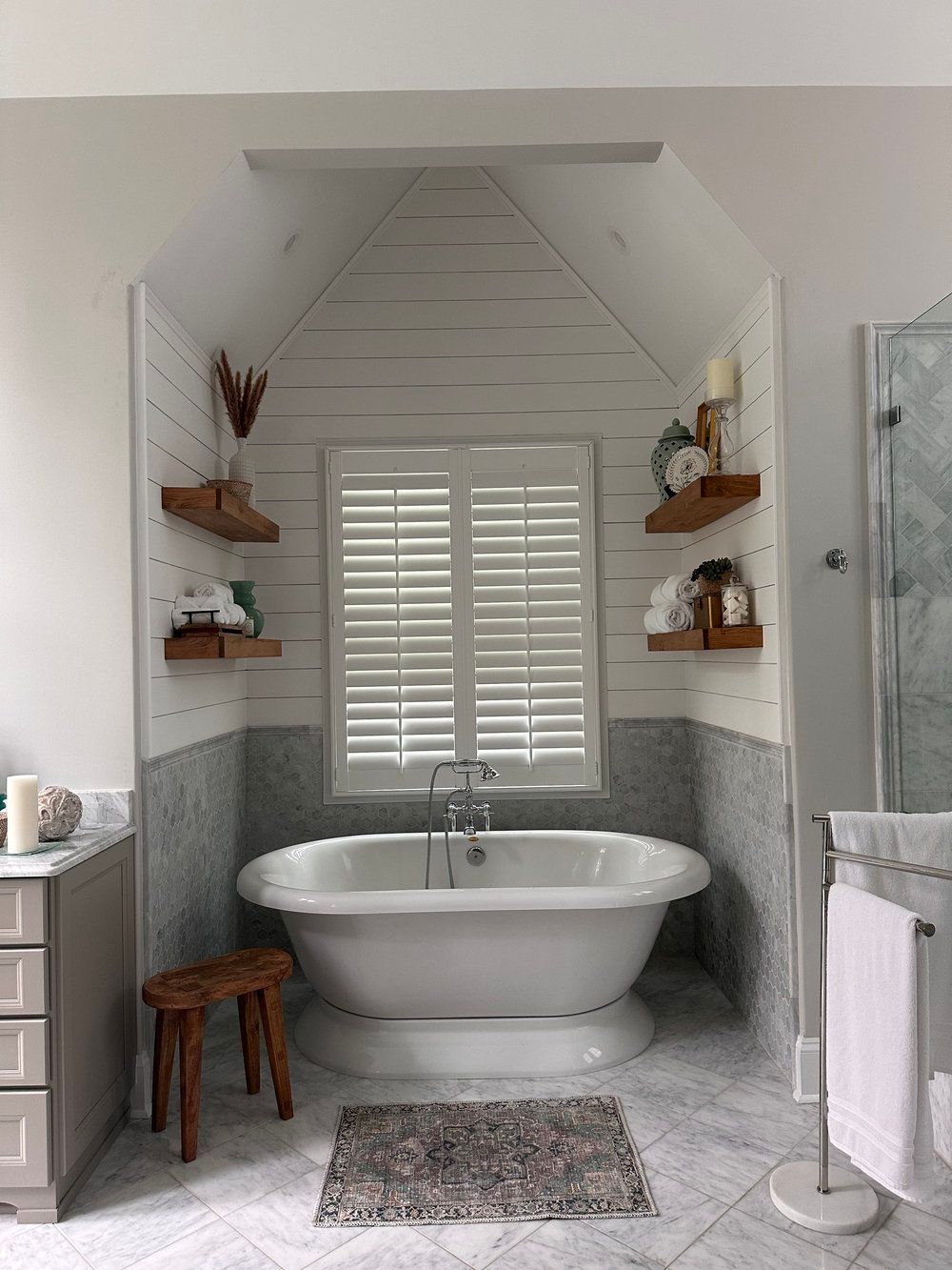 Bright bathroom with a clawfoot tub, white shutters, gray wainscoting, shelves, and a stool.