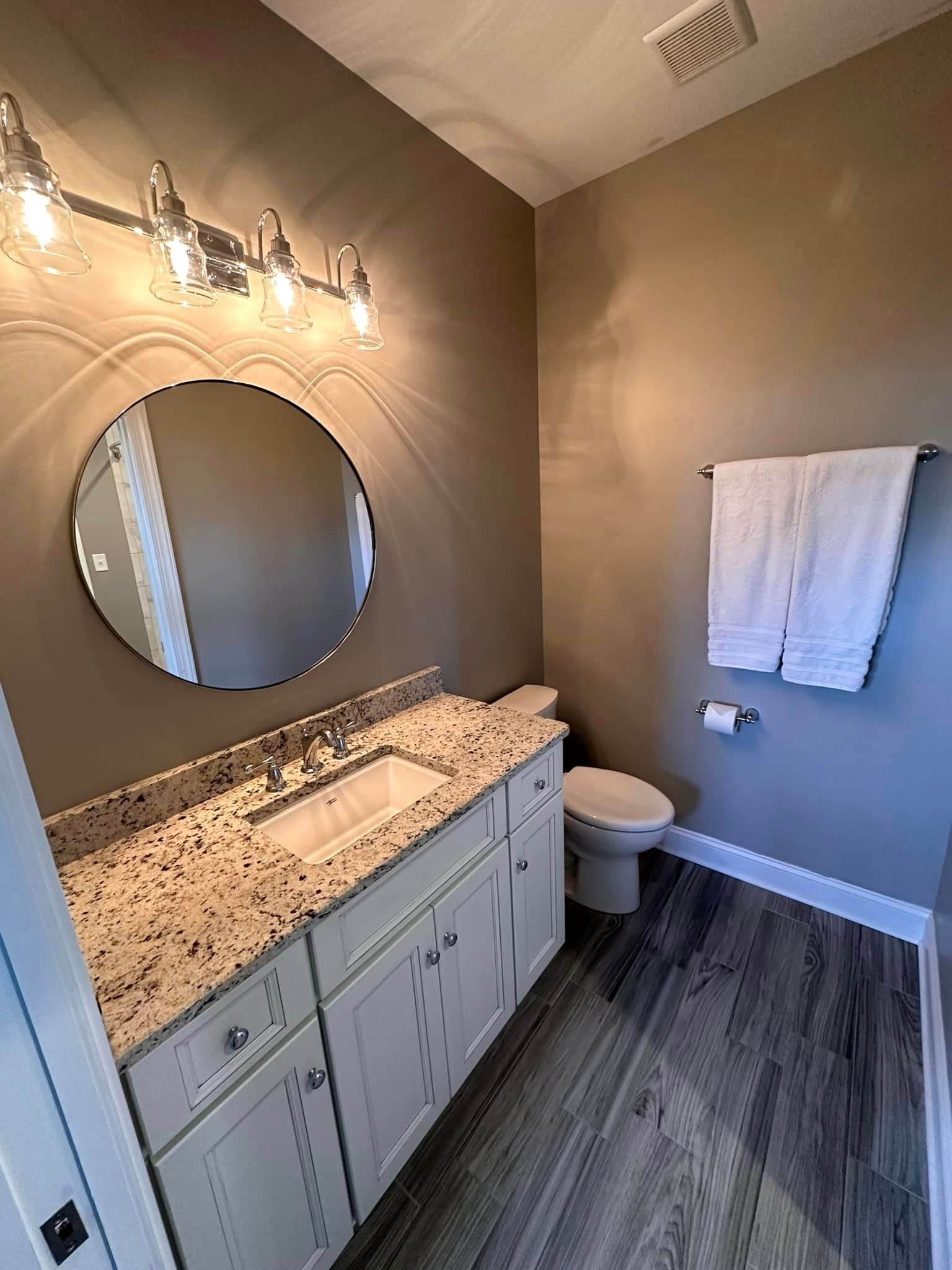 Small beige bathroom with granite vanity, round mirror, towel rack, toilet, and gray wood-look floor.