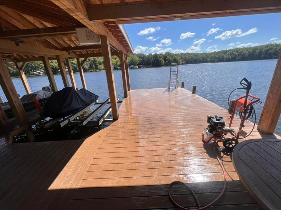 Covered lakeside dock with wooden deck, lounge chairs, fishing gear, and calm water under a bright blue sky