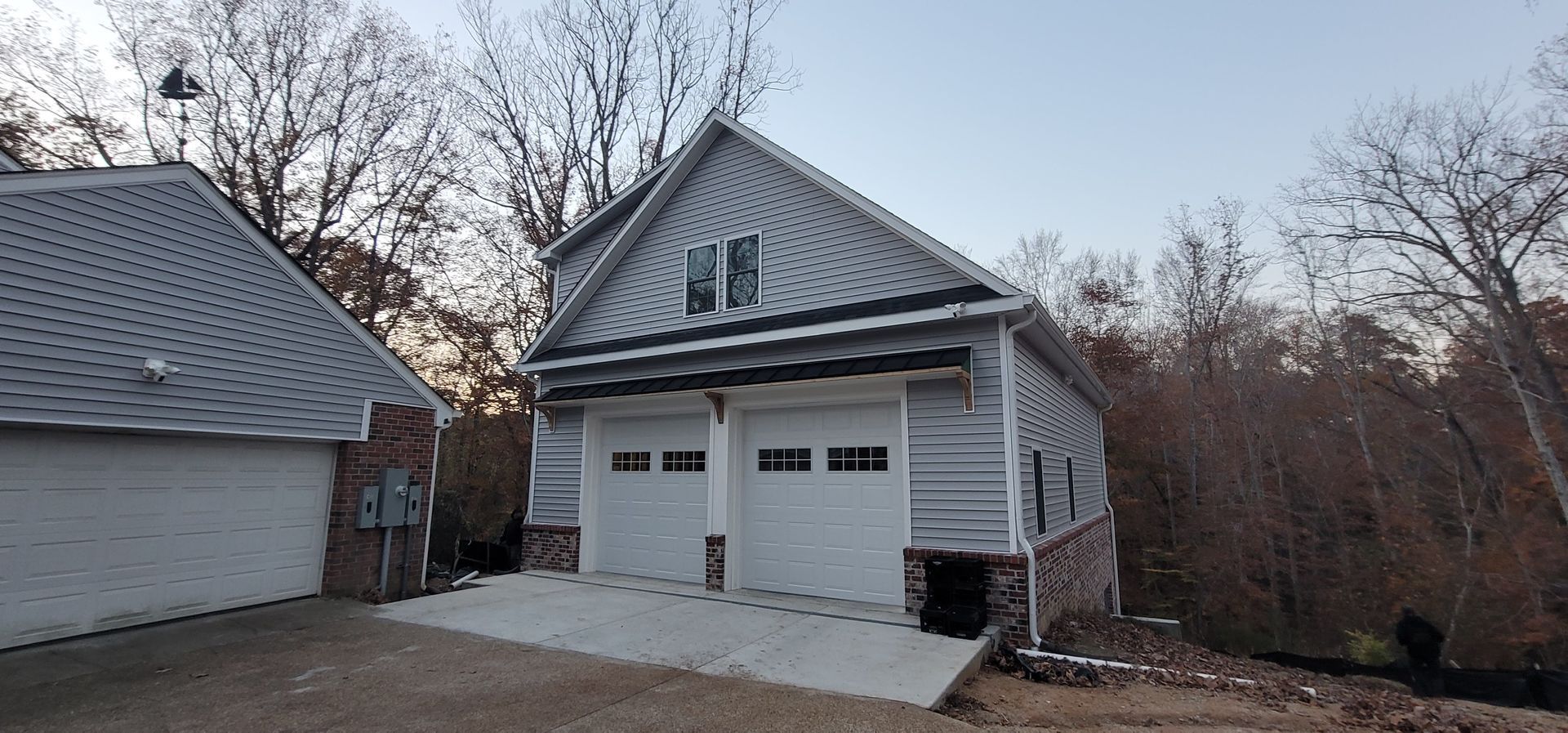 Detached white garage with two doors and a steep metal roof beside a house in a wooded yard