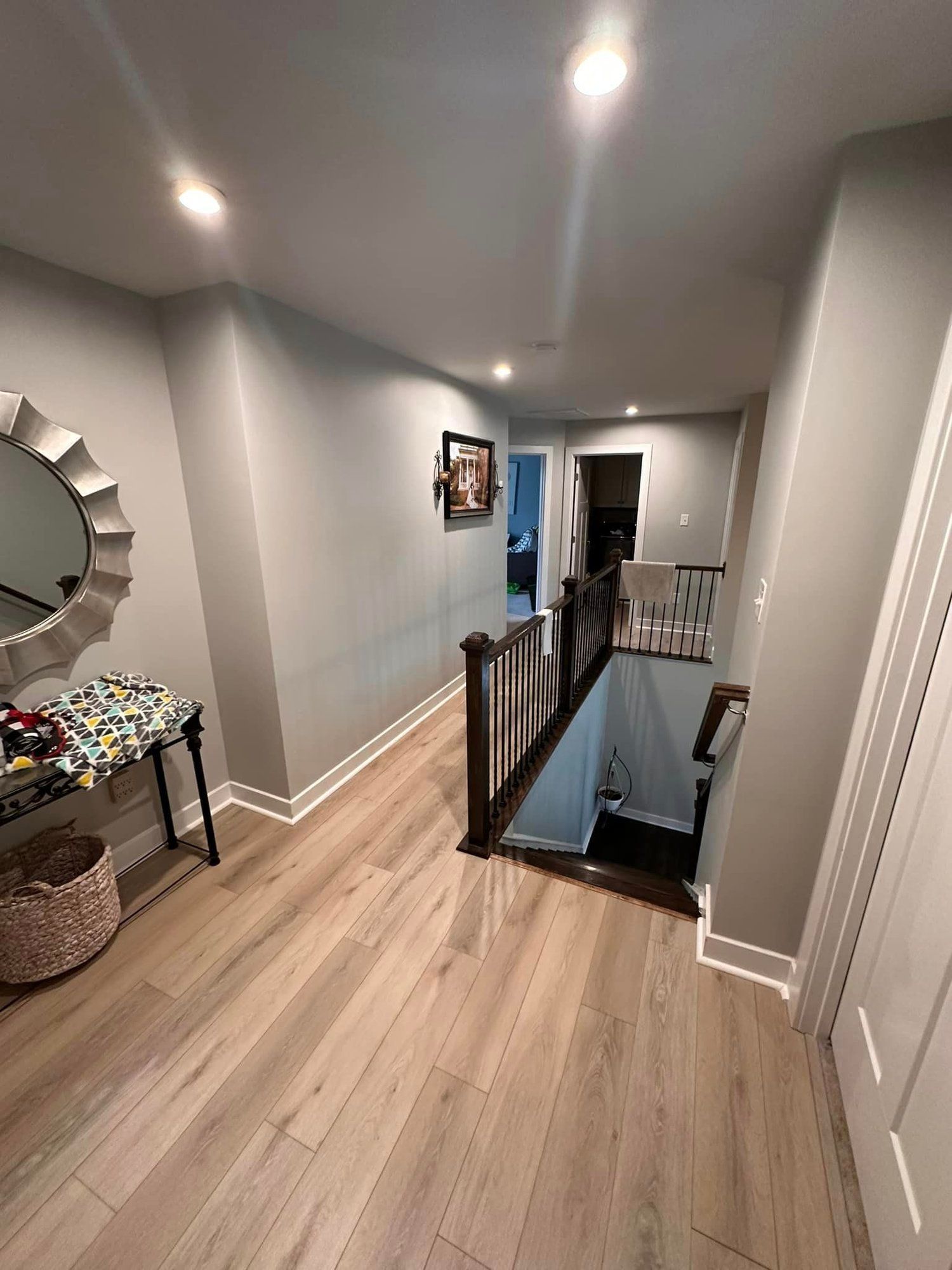 Narrow hallway with gray walls, wood floors, mirror, and a staircase opening with black railing.