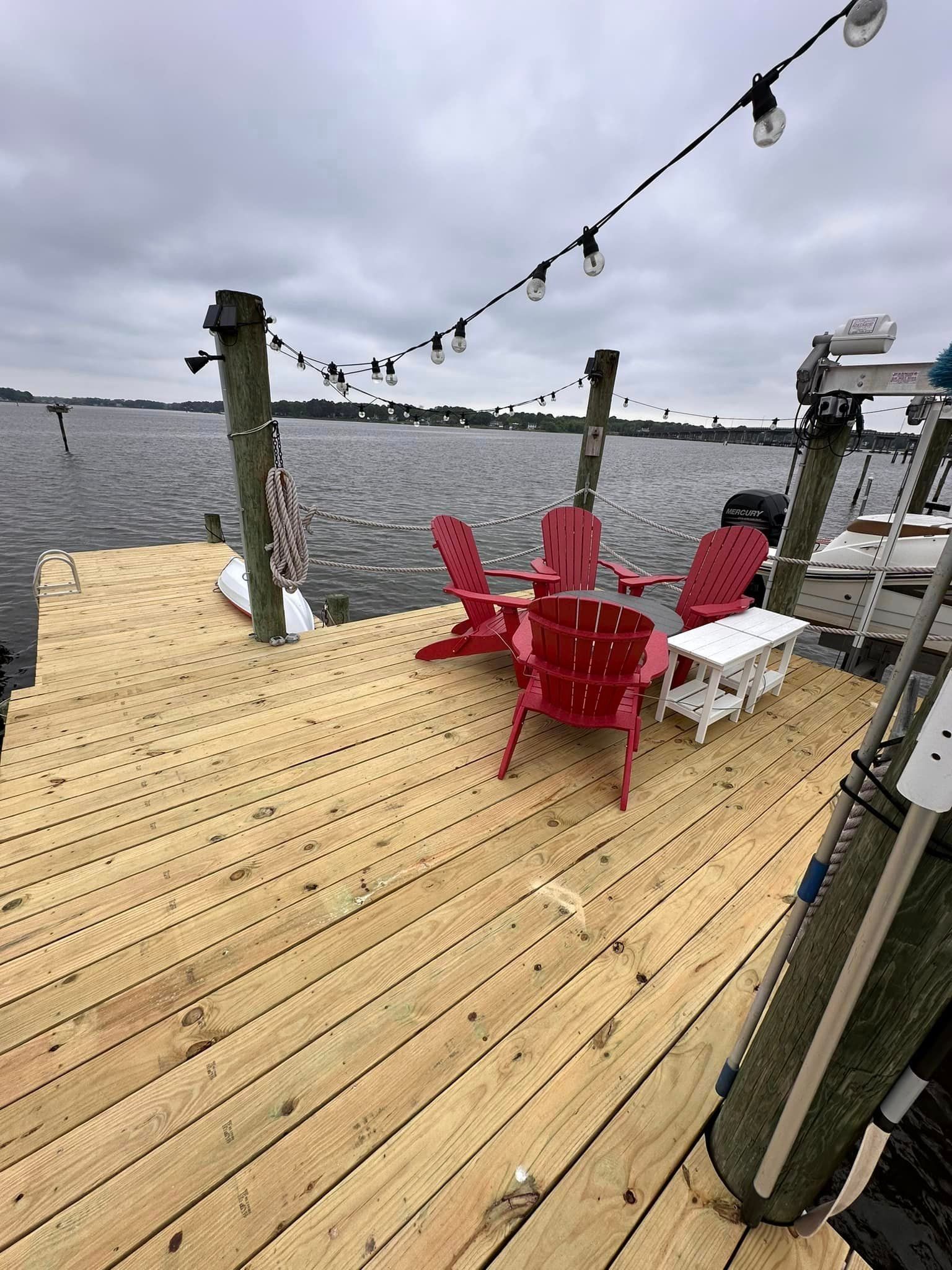 Wooden dock by the water with red Adirondack chairs, white loungers, and string lights under a cloudy sky