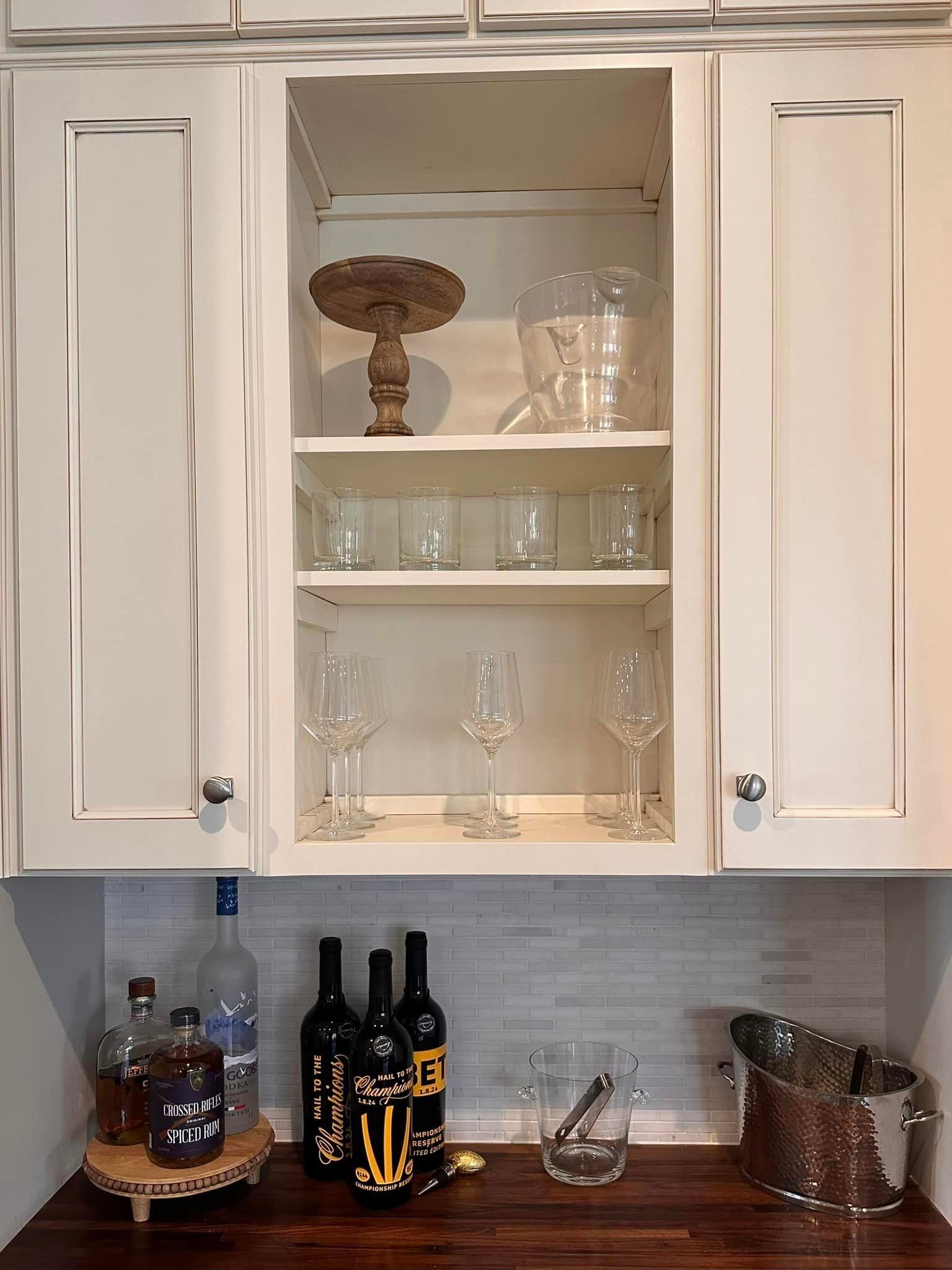 White kitchen cabinets with glassware, bottles, and bar tools on a countertop in a home bar setup