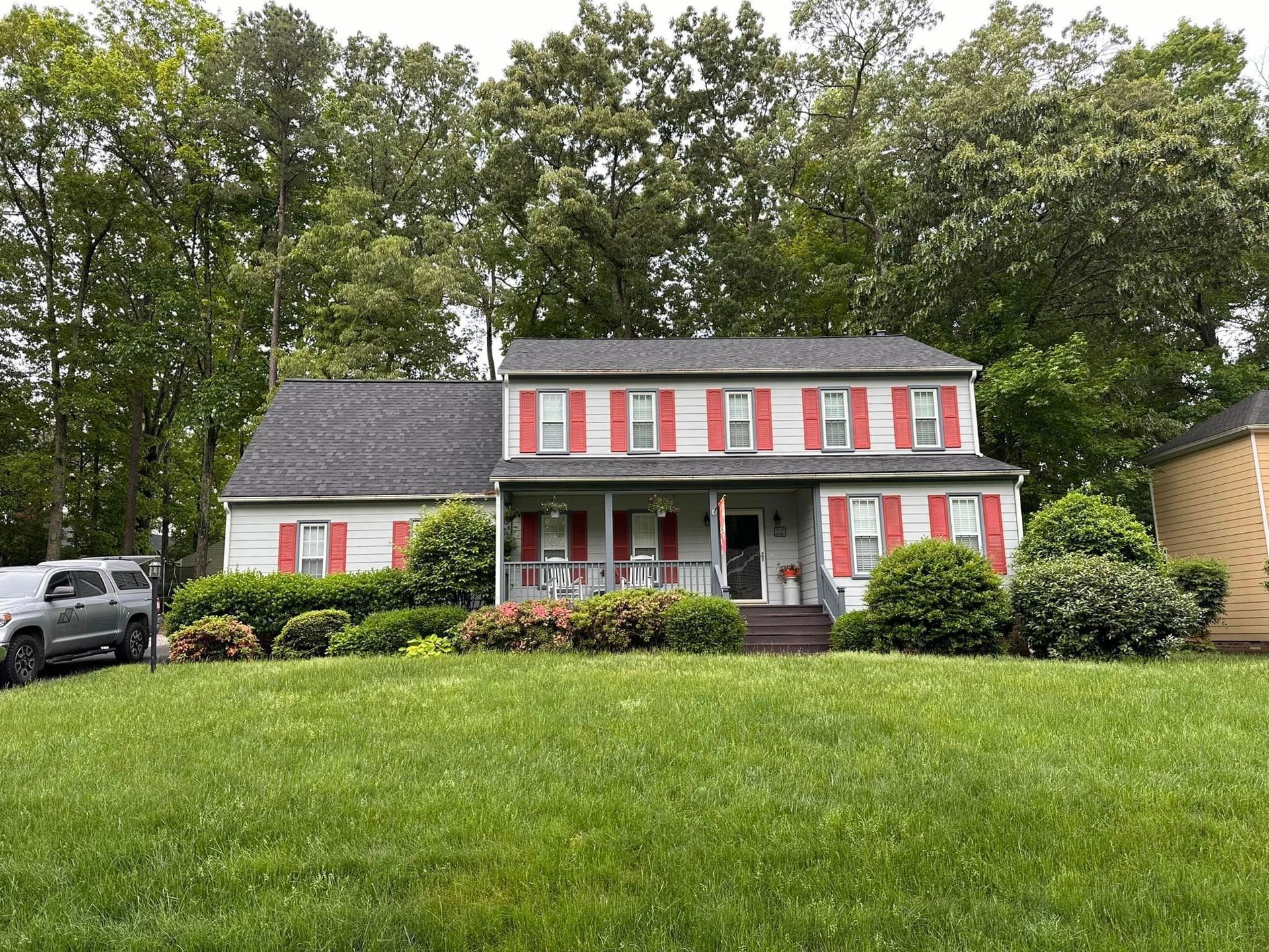Two-story white house with red shutters and porch, set behind a green lawn and trees