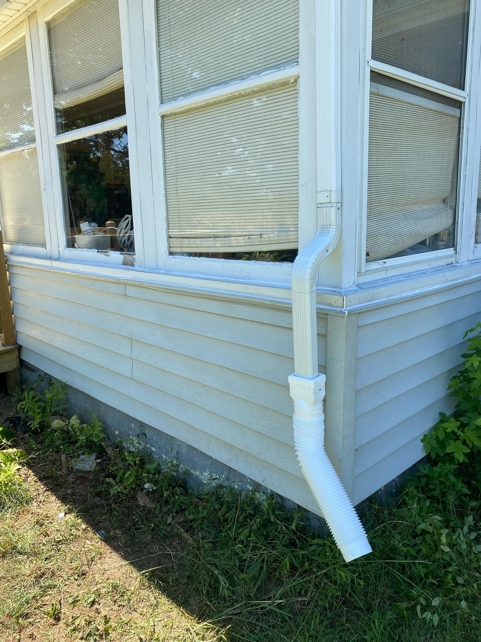 White house corner with bay windows and a white downspout over vinyl siding and grass below