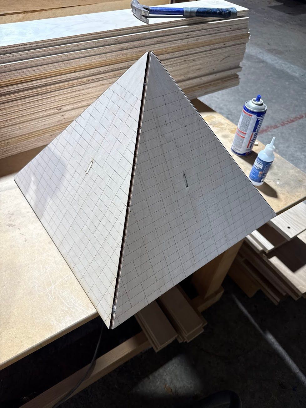 White pyramid model on a workbench, with stacked wood and spray cans in the background.