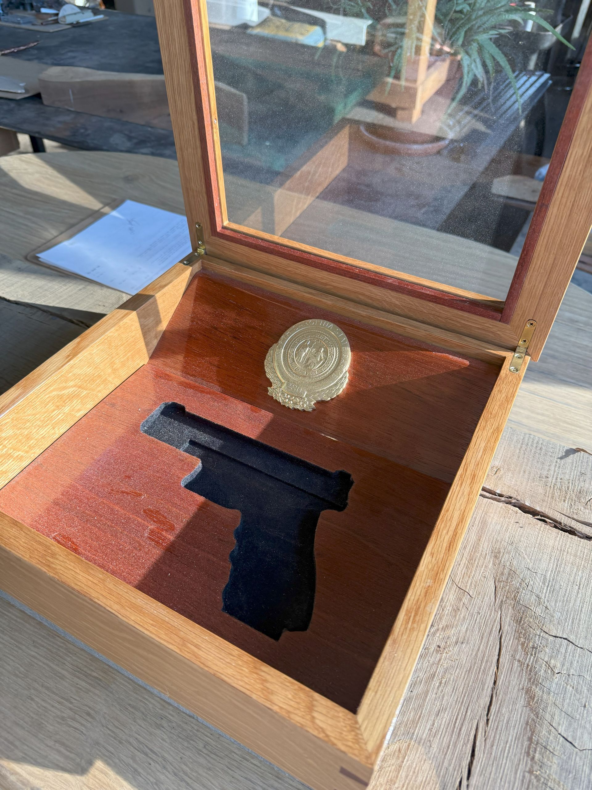 Open wooden display box with a black handgun and a round gold-colored object inside on a patio.