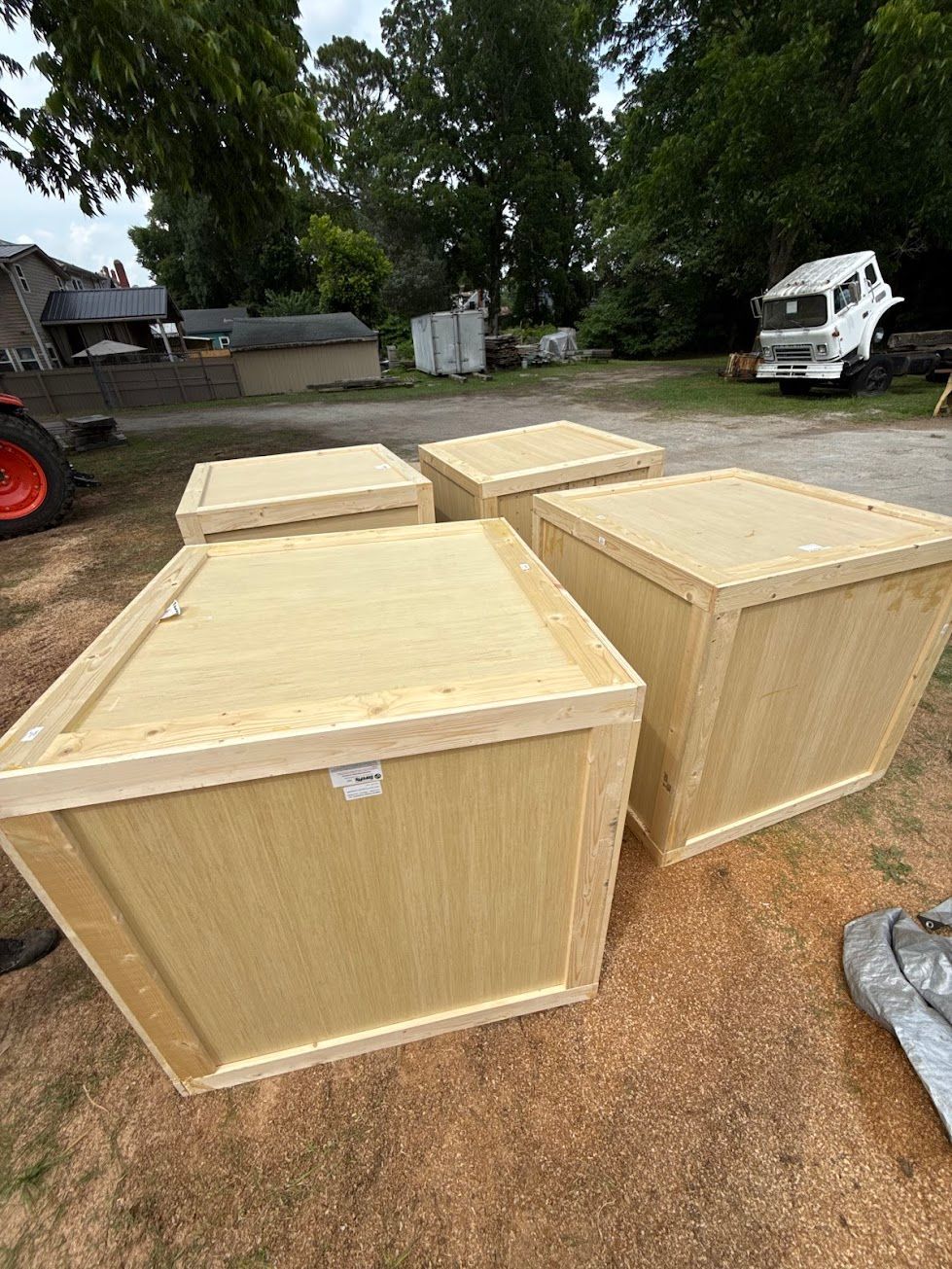 Four large wooden crates outdoors on a dirt lot, with trees, a truck, and an RV in the background