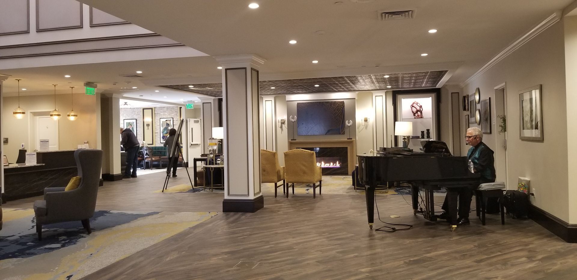 A man plays a grand piano in a hotel lobby.  Fireplace and seating are in the background.