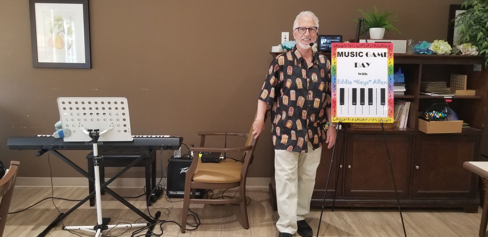 Man standing beside a music stand and keyboard in a room, with a piano and cabinets behind him.