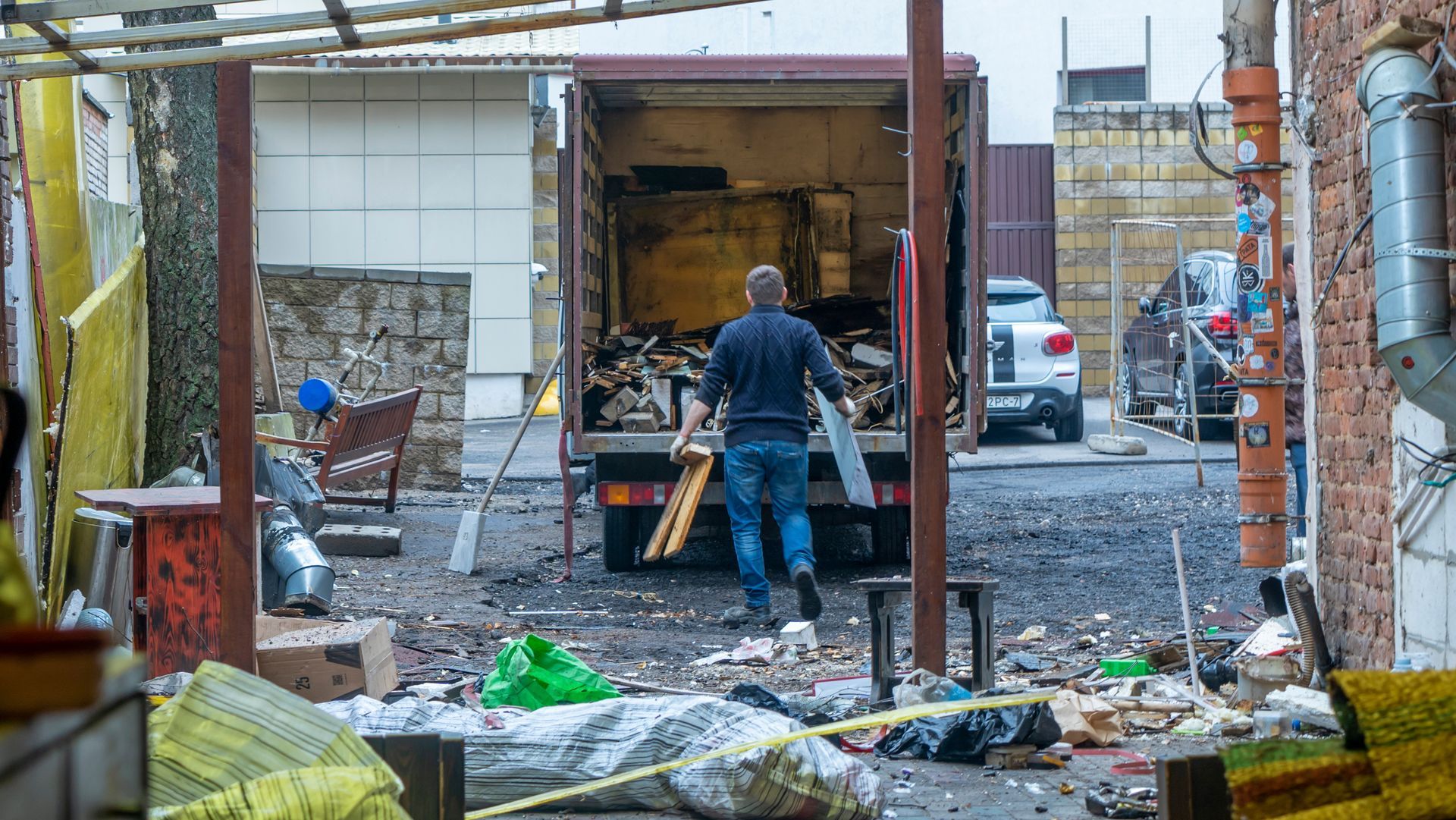 A person in a blue shirt walks toward an open truck parked in an urban area filled with scattered debris and trash.
