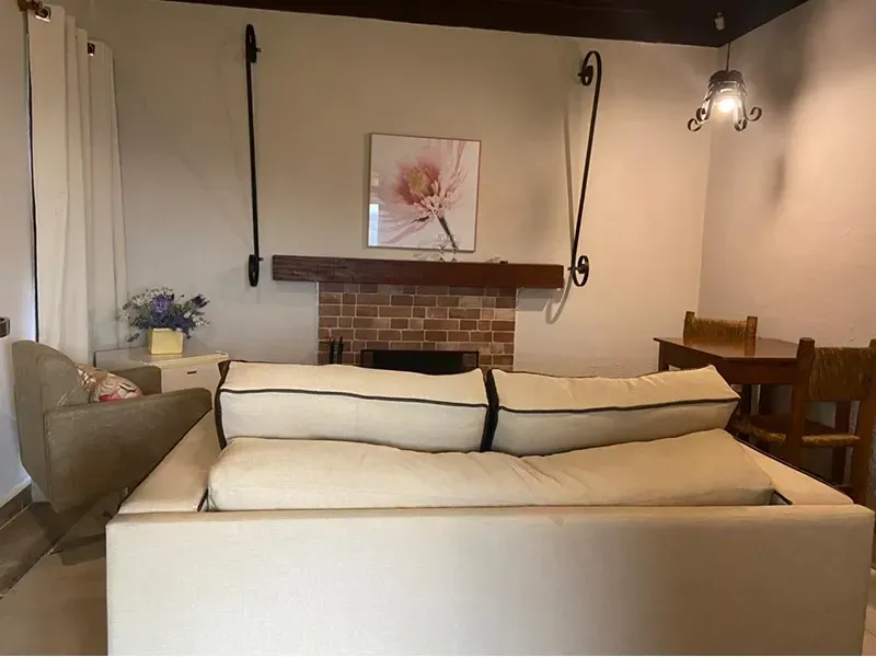 Sala de estar com um sofá bege claro em primeiro plano, uma lareira de tijolos, uma estampa floral na parede e um pequeno conjunto de jantar.