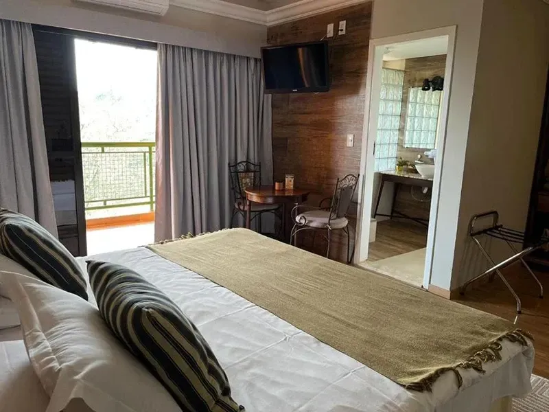 Um quarto de hotel com uma cama com peseira bege, parede com painéis de madeira, duas cadeiras e uma porta de banheiro aberta.