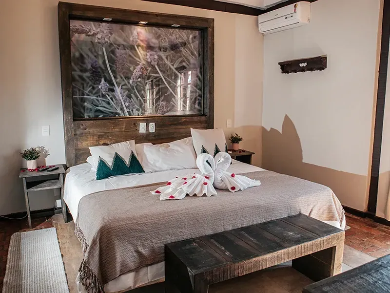 Um quarto de hotel com a cama arrumada, decorada com cisnes feitos de toalhas, uma grande obra de arte com temática da natureza na parede e um banco de madeira aos pés da cama.