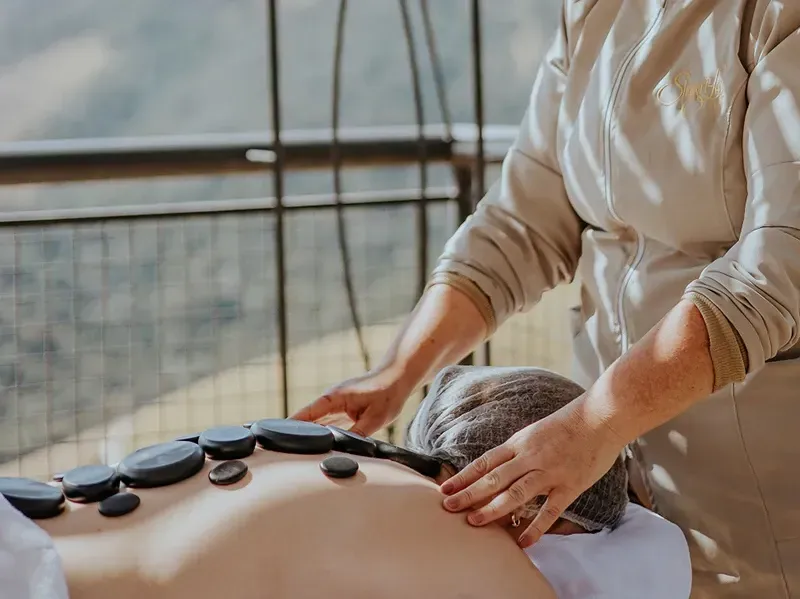 Uma pessoa recebe uma massagem com pedras quentes nas costas enquanto está deitada perto de uma varanda com uma vista panorâmica da montanha.