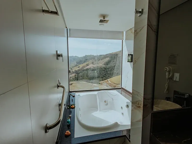 Um banheiro de hotel moderno com uma banheira de imersão branca ao lado de uma grande janela do chão ao teto com vista panorâmica para as montanhas.