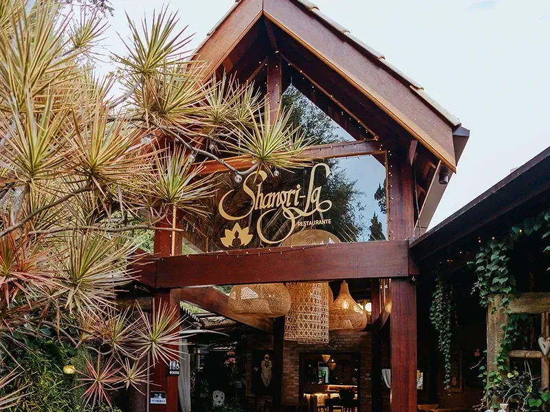 A entrada de madeira para os Jardins Shangri-La, com uma placa de vidro, luminárias de vime penduradas e plantas exuberantes ao redor.