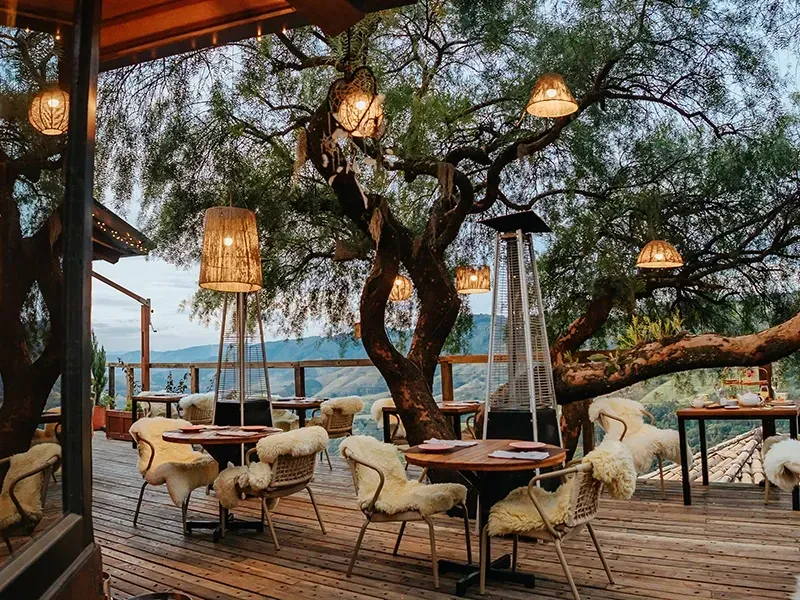 Pátio externo do restaurante com mesas, cadeiras revestidas de pele de carneiro e lanternas penduradas sob grandes árvores ao entardecer.