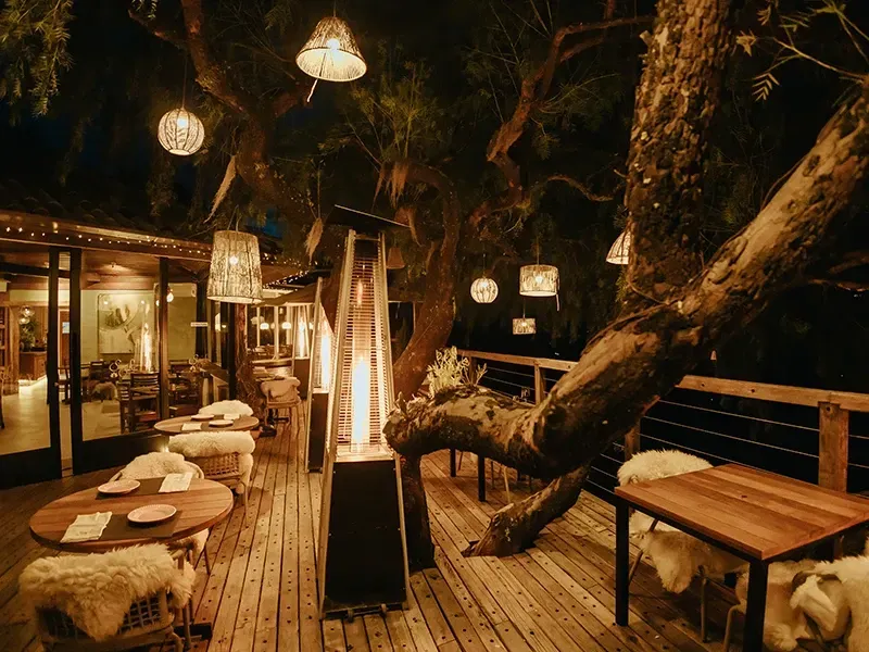 Restaurante com deck externo à noite, com mesas de madeira, cadeiras aconchegantes de pele de carneiro, luzes suspensas e um aquecedor central no pátio.