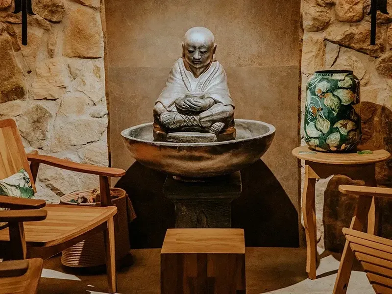 Uma estátua de monge feita de pedra está situada em uma fonte, entre duas cadeiras de madeira, encostada em uma parede de pedra texturizada, em uma sala com pouca luz.