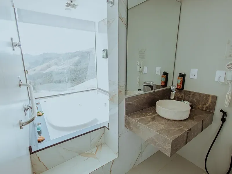 Um banheiro moderno com uma banheira de imersão branca junto a uma janela com vista para a montanha, uma bancada de mármore e uma pia circular.