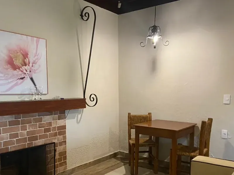 Uma lareira de tijolos com uma estampa floral rosa acima dela, perto de uma pequena mesa de madeira com duas cadeiras sob uma luminária pendente.