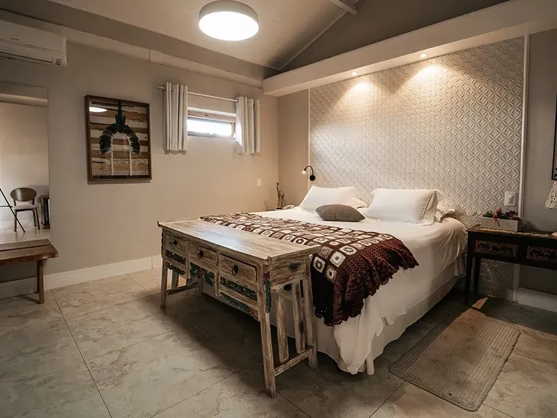 Um quarto com cama de casal com um banco de madeira aos pés, cabeceira estampada e piso de azulejo claro.