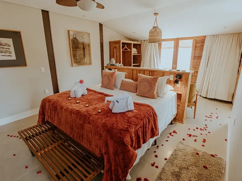 Um quarto romântico com uma cama coberta por um cobertor vermelho-alaranjado, dois cisnes feitos de toalhas e pétalas de rosas vermelhas espalhadas.