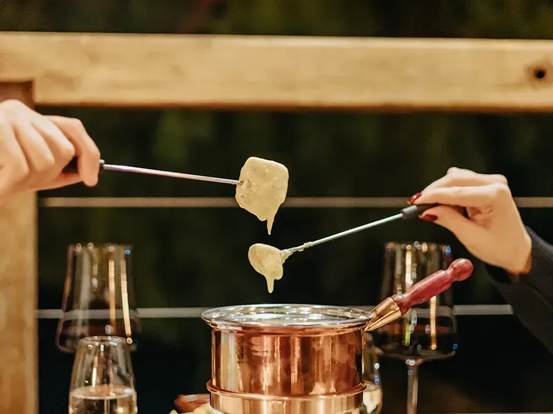 Duas pessoas usam garfos compridos para mergulhar o pão em uma panela de cobre para fondue, que está sobre uma mesa com taças de vinho.