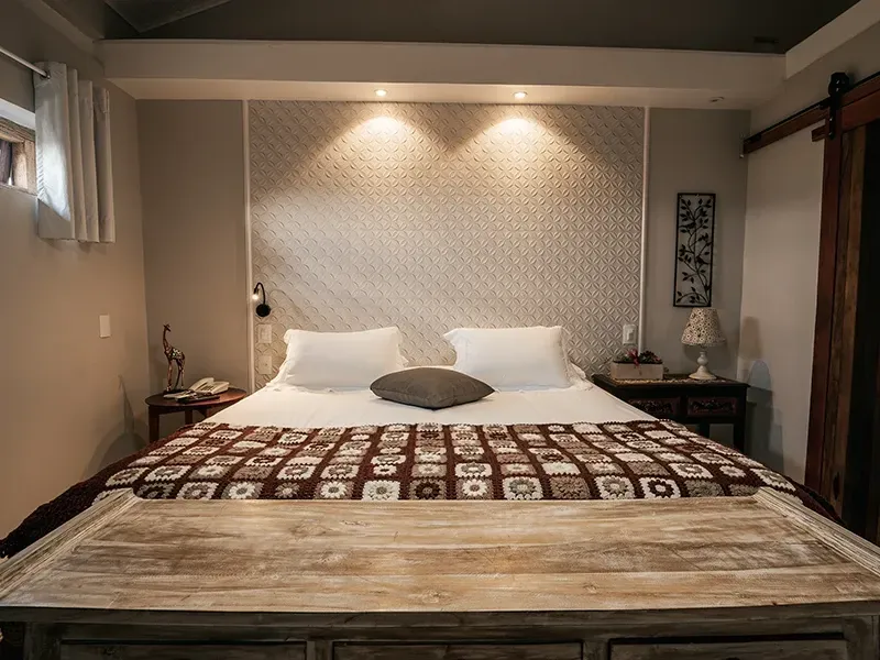 Uma cama king-size com colcha de estampa geométrica, travesseiros brancos e um banco rústico de madeira em um quarto de hotel com iluminação suave.