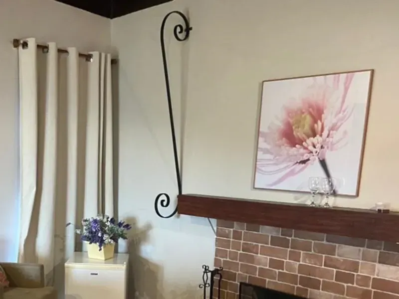 Um quarto com cortinas leves, uma pequena mesa com flores, uma lareira de tijolos e um alto gancho decorativo preto em forma de pergaminho na parede.