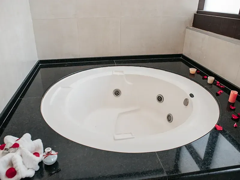 Uma banheira de hidromassagem branca e redonda, embutida em uma bancada de granito preto, decorada com pétalas de rosa, velas e toalhas dobradas.