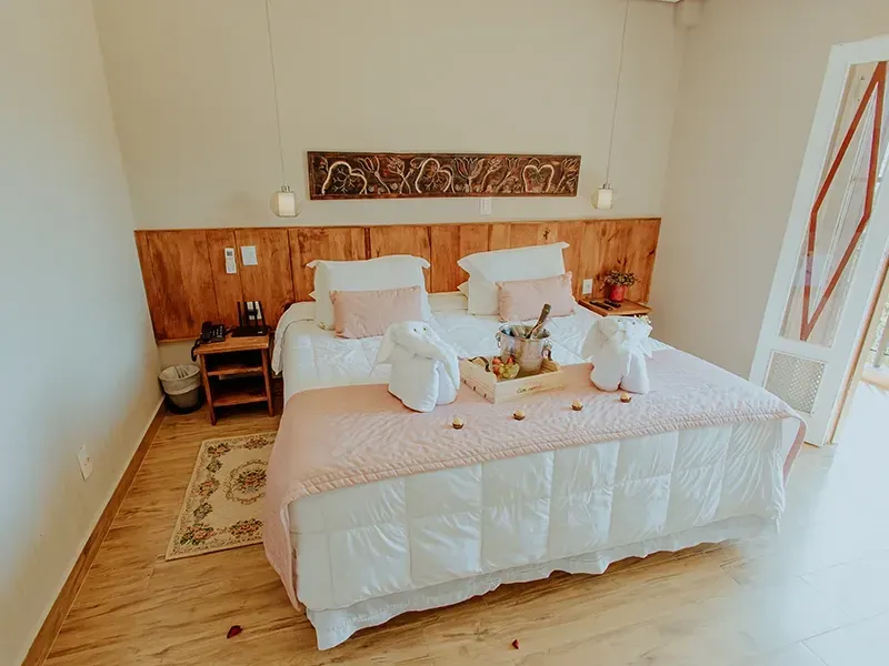 Um quarto de hotel aconchegante com cama de cabeceira de madeira, roupa de cama branca, dois bichinhos de toalha e uma bandeja de café da manhã.