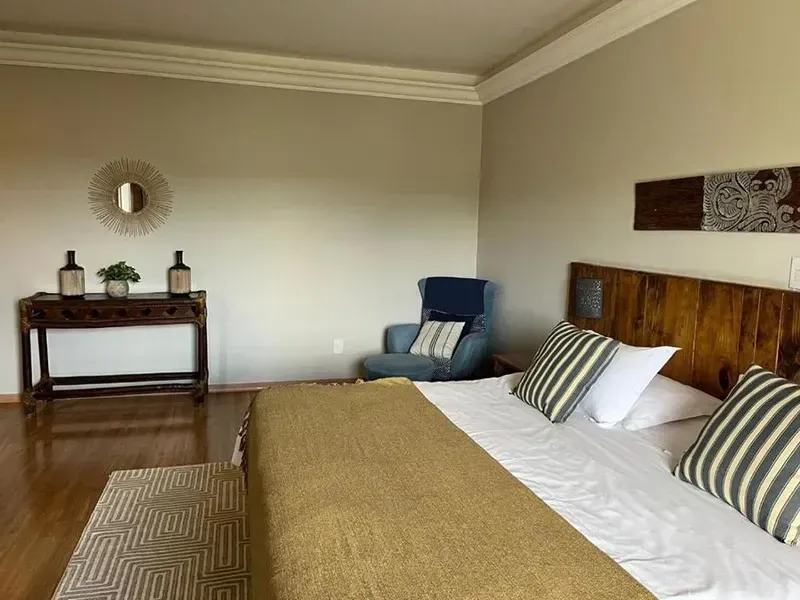 Um quarto com uma cama com uma manta dourada, almofadas listradas, uma cabeceira de madeira, uma poltrona azul e uma mesa de apoio.