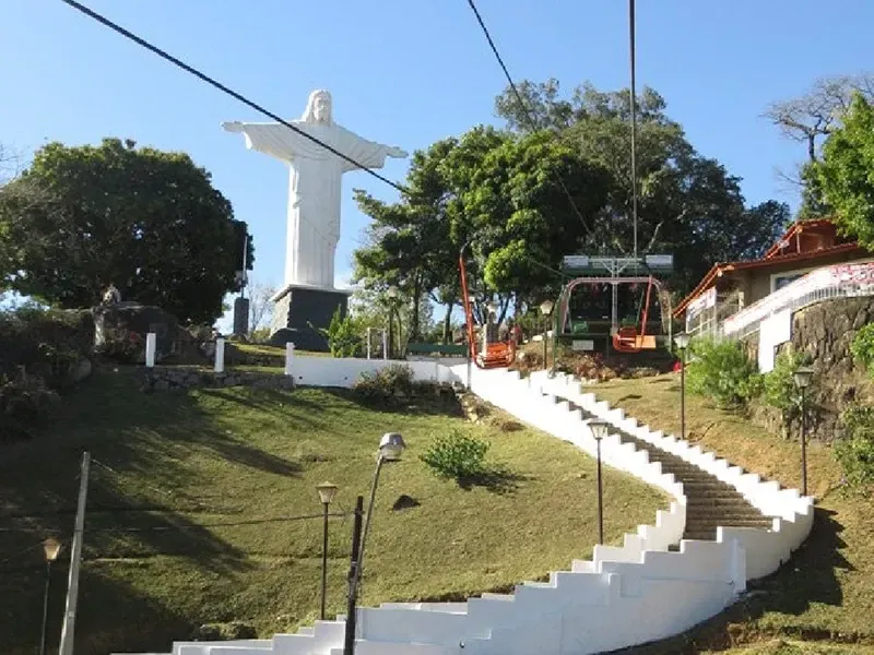 Uma estátua branca de Cristo ergue-se numa colina acima de uma escadaria em espiral, com cabos de teleférico acima, contra um céu azul.