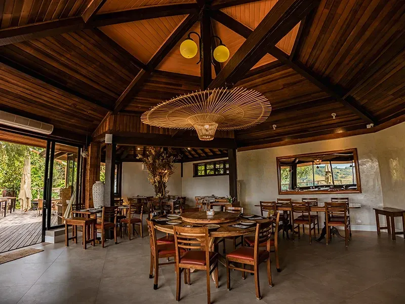 Uma sala de jantar com teto rústico de madeira, um grande lustre pendente de vime, mesas redondas e cadeiras de madeira.