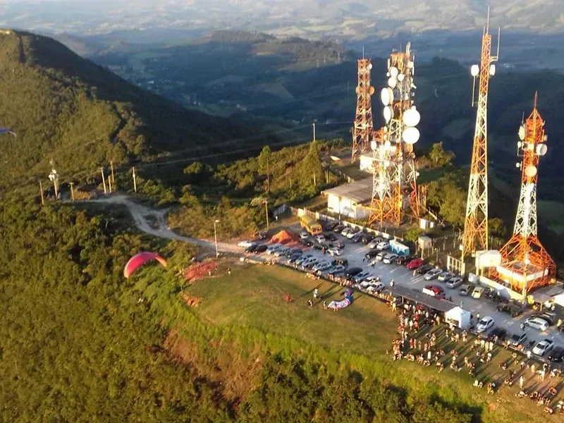 Vista aérea de um pico de montanha com torres de transmissão, um estacionamento e um parapente voando próximo à encosta gramada.