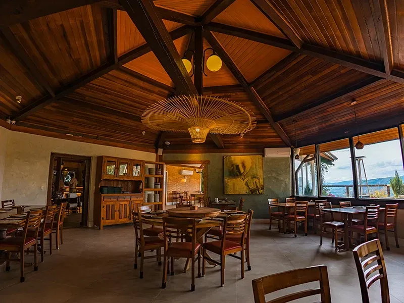Uma sala de jantar rústica com tetos abobadados de madeira, iluminação aconchegante e mesas dispostas perto de grandes janelas com vista.