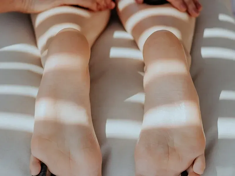 Uma pessoa está deitada de bruços, com os pés descalços apoiados em uma maca de massagem, iluminada pela luz do sol que filtra através das persianas.