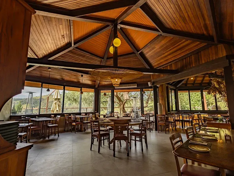 Interior de restaurante rústico com vigas de madeira no teto, luminárias pendentes e mesas dispostas perto de grandes janelas com vista para o mar.