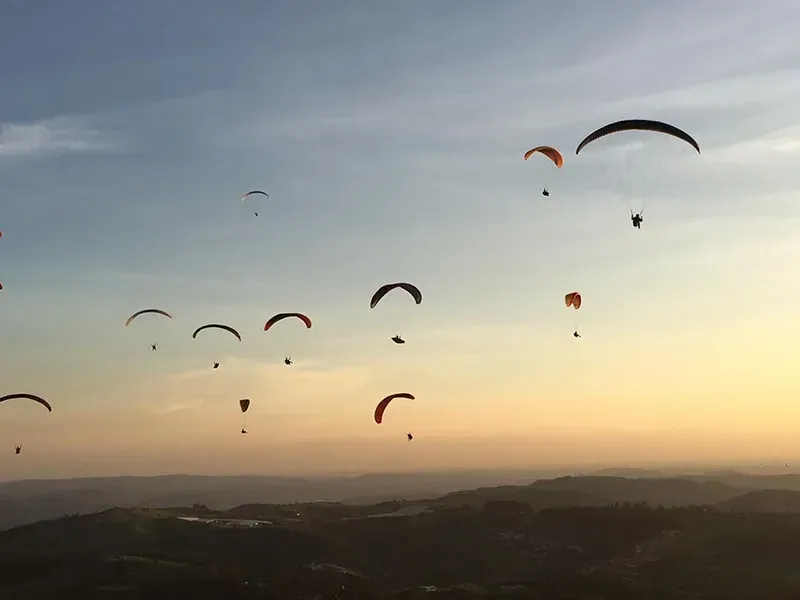 Vários parapentes planam em um céu dourado e enevoado sobre uma paisagem montanhosa ao pôr do sol.